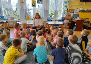Dzieci siedzą na dywanie słuchają opowiadania o wróbelku Elemelku czytanego przez nauczycielekę.
